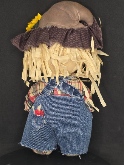 Backside of a handmade fabric scarecrow doll with a brown head, beige straw hair, a dark purple knit hat, and blue denim shorts, laid flat on a black background.