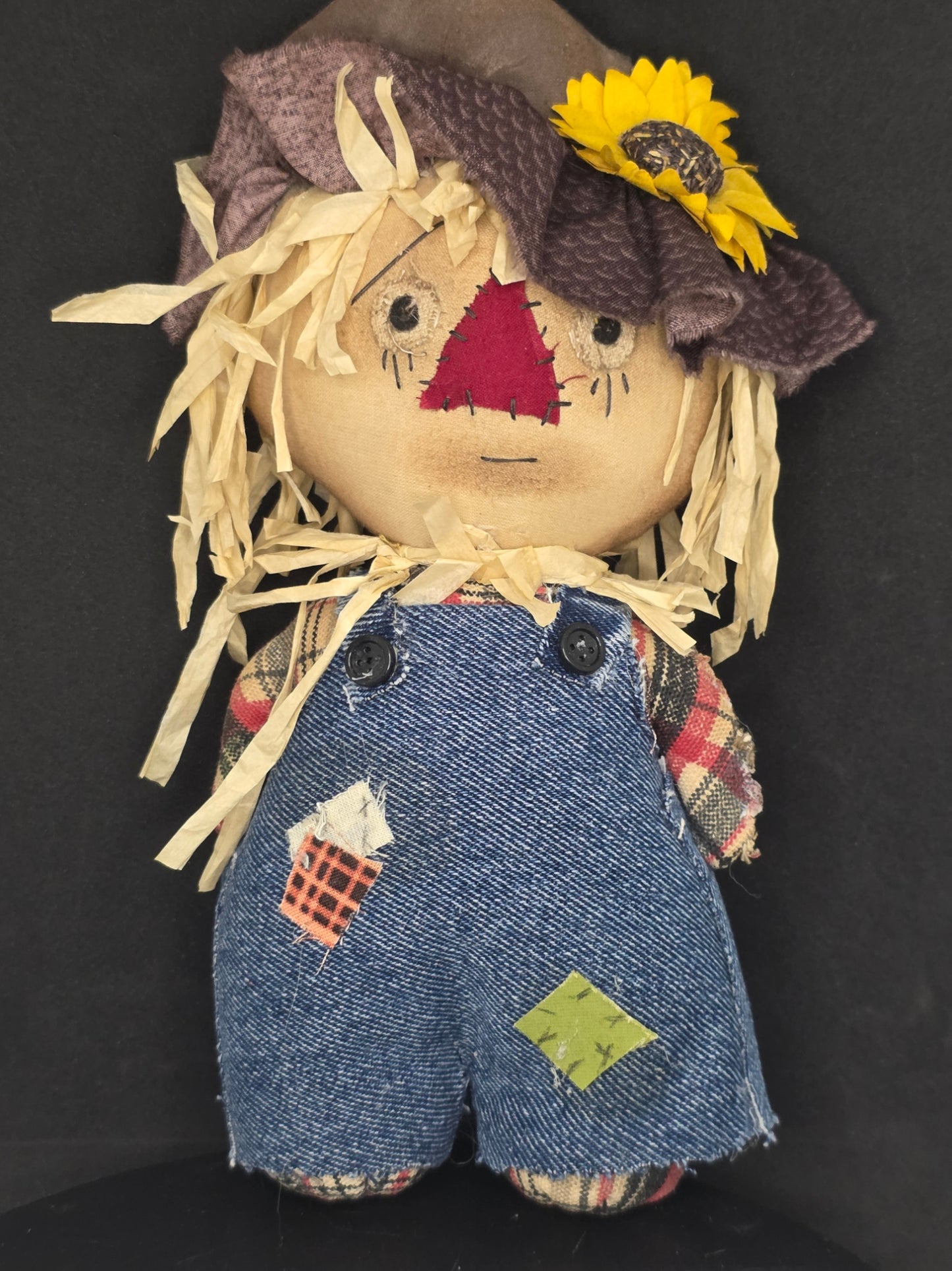 A handmade fabric scarecrow doll with a beige face, yellow straw hair, a brown knit hat with a yellow sunflower, and blue denim-style overalls with patchwork details, shown on a black background.