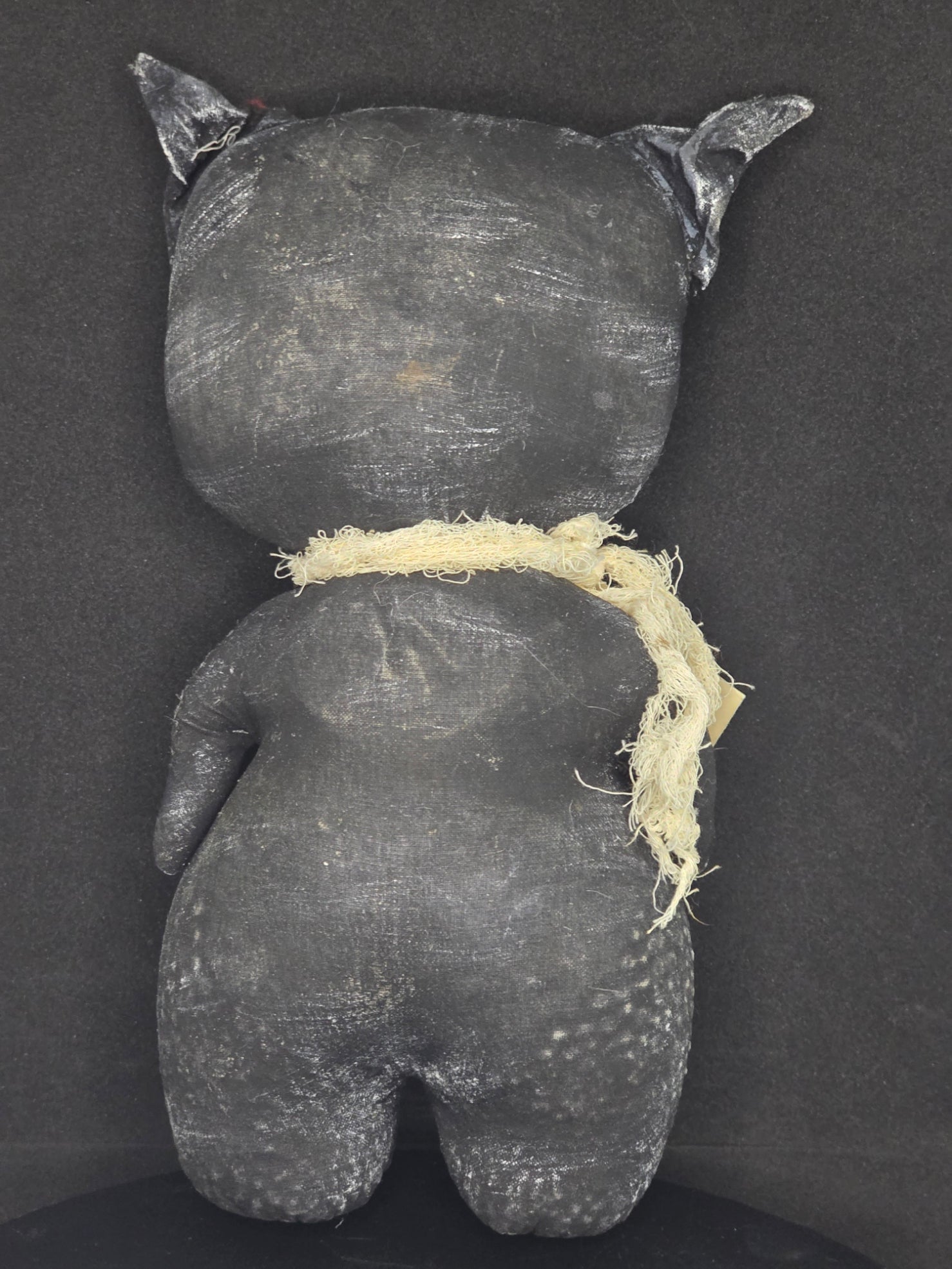 Backside of handmade black cat doll photographed from above on a dark fabric background. A frayed off-white band runs around the center.