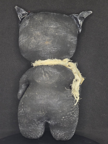 Backside of handmade black cat doll photographed from above on a dark fabric background. A frayed off-white band runs around the center.