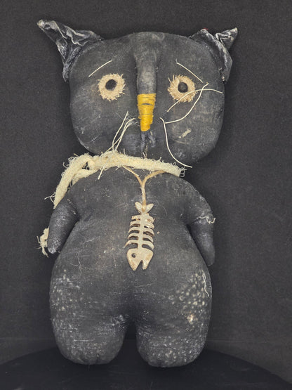A distressed black fabric cat doll with embroidered white eyes, a yellow nose, and a white fish bone necklace, lying on a dark background.