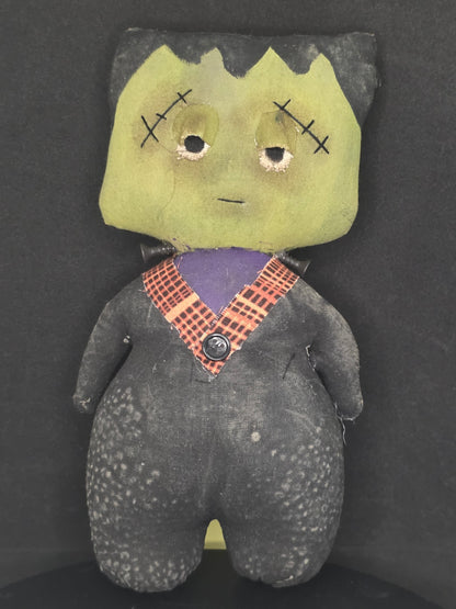 A handmade Frankenstein doll with a stitched green face, black hair, and a purple and orange plaid shirt, lying on a dark background.