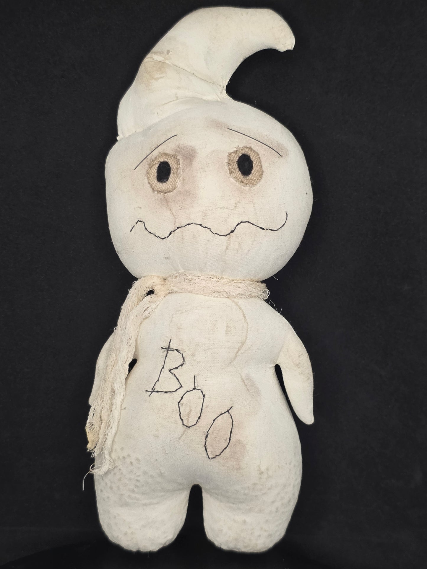 A white fabric doll shaped like a ghost or elephant with embroidered eyes, stitched facial features, and the word 'BOO' written on its side, photographed against a black background.