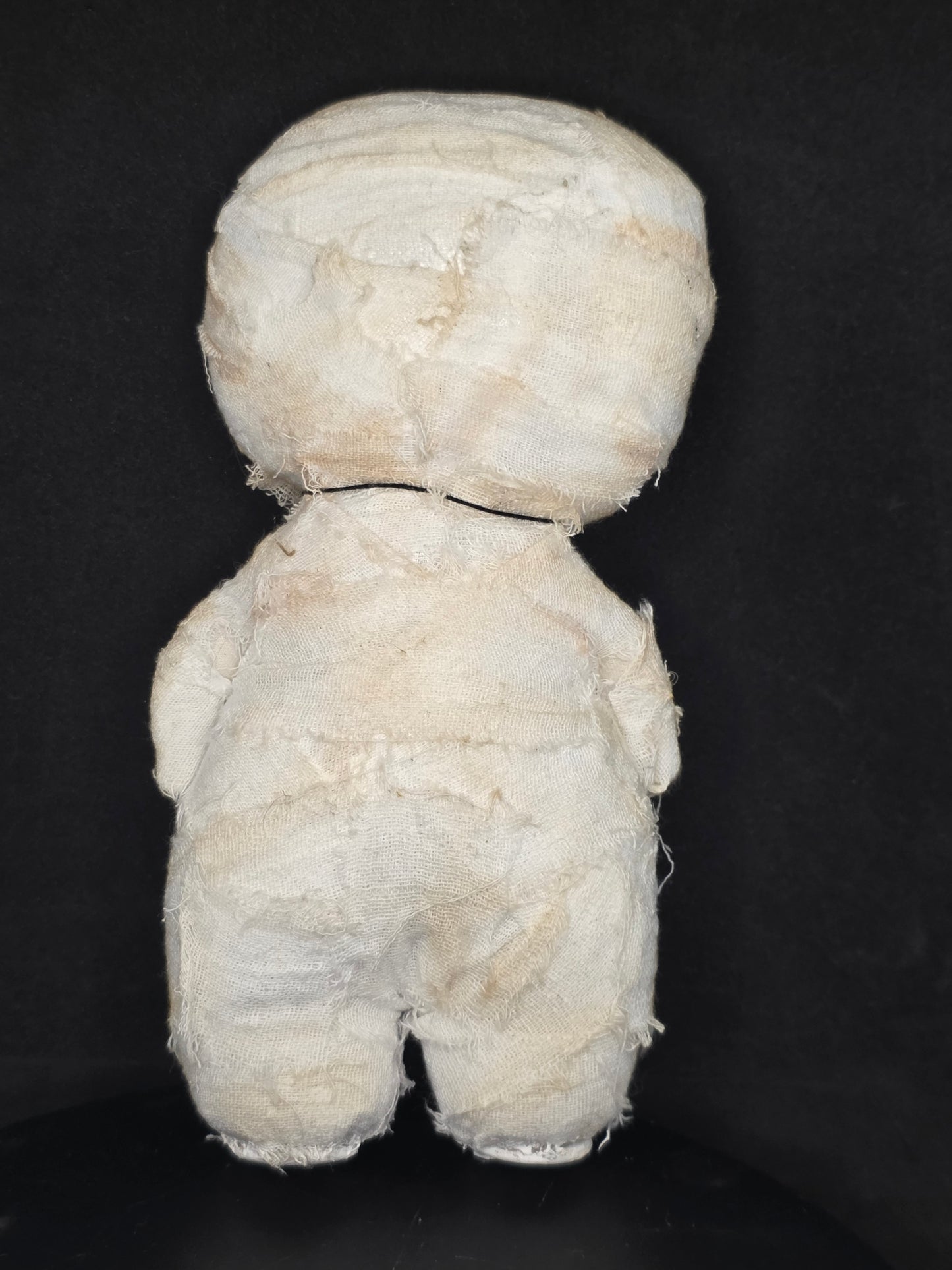 Backside of a handmade mummy doll wrapped in off-white gauze or cloth, photographed from above on a dark black background.