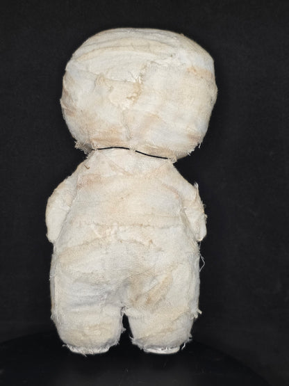 Backside of a handmade mummy doll wrapped in off-white gauze or cloth, photographed from above on a dark black background.