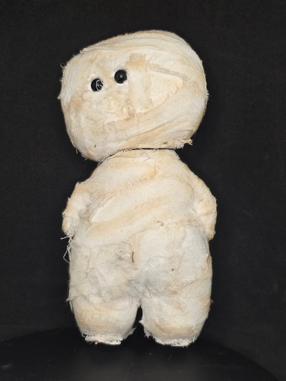 A mummy doll wrapped in off-white fabric, with small black bead eyes, photographed on a dark black background.