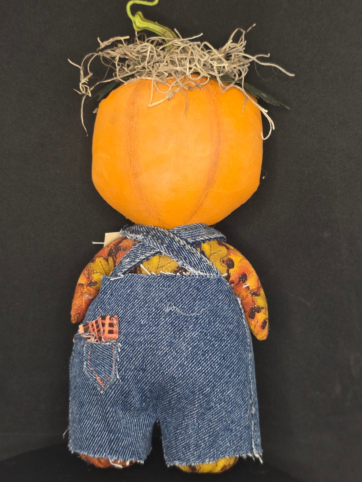 Backside of a handmade rag doll pumpkin with a bright orange body, denim blue fabric pants, and a patterned yellow and brown shirt, lying on a black background.