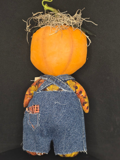 Backside of a handmade rag doll pumpkin with a bright orange body, denim blue fabric pants, and a patterned yellow and brown shirt, lying on a black background.