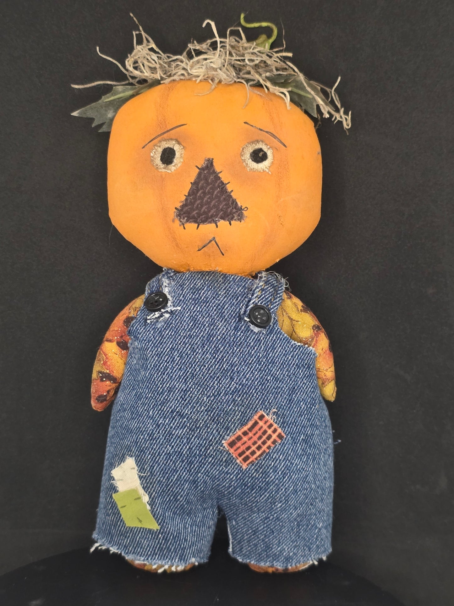 A handmade doll figure with a round orange pumpkin head, stitched facial features, a bundle of straw hair, and blue denim shorts with patchwork fabric details, photographed on a dark background.