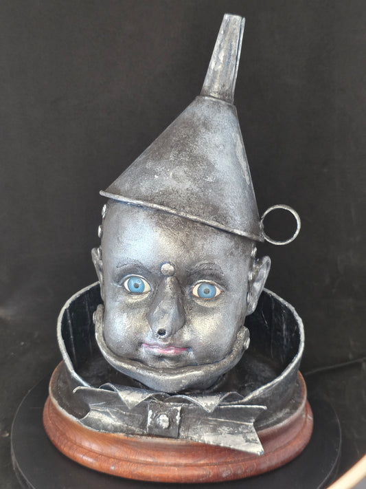 Wizard of Oz Tin Man Bust original mixed media sculpture — porcelain doll face painted in aged metallic silver with blue glass eyes, hand-crafted tin funnel hat with ring detail, metal-like collar and bow tie, mounted on a warm wood base against a dark background