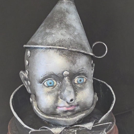 360-degree view of the Wizard of Oz Tin Man Bust sculpture, showing the aged metallic silver porcelain face, blue glass eyes, tin funnel hat, metal-like collar and bow tie details, and warm wood base from all angles