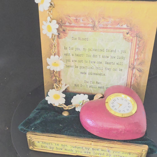 360-degree view of the Wizard of Oz Tin Man's Heart working clock and display stand, showing the heart clock, stainless steel chain with daisy accents, gold metallic acrylic stand with dimensional movie quote plaque, and dark green velvet cushion base