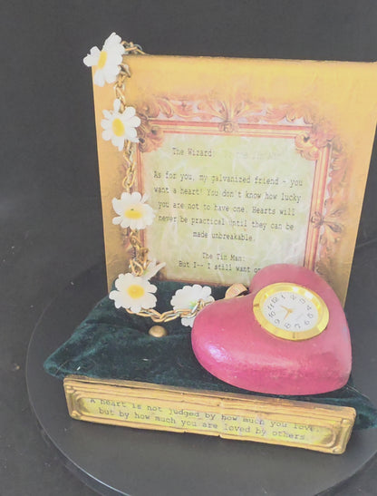 360-degree view of the Wizard of Oz Tin Man's Heart working clock and display stand, showing the heart clock, stainless steel chain with daisy accents, gold metallic acrylic stand with dimensional movie quote plaque, and dark green velvet cushion base